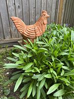 Side view of terracotta rooster statue partially surrounded by green foliage, showing textured feather details and tail.