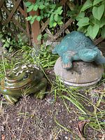 Frog and turtle statues outdoors in garden area with plants and lattice fencing behind. Turtle is on concrete stand. Frog has worn paint.