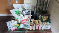 Photo showing multiple gardening products including large white bags of Lawn Fertilizer and Moss Control, Rose Food, and other fertilizers, alongside bottles of plant treatment liquids and small boxed plant food products all lined on the floor.