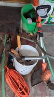 Photo shows Black & Decker electric weed whacker handle, green garden spreader and hand gardening tools in a white bucket with orange extension cords coiled nearby.