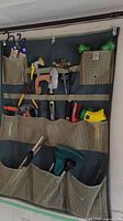 Canvas wall organizer hanging, showing multiple gardening tools in pockets: hand saw, pruning shears, sprayer, clippers, trowels.