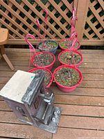 Six red hanging planters with soil and some plant remnants on a wooden deck beside a rustic wooden birdhouse with hinged roof and perch.