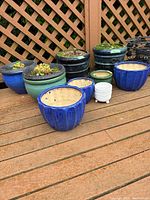 Nine ceramic pots arranged on deck against lattice, including blue, green, black and white pots.
