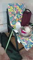 Photo of four green plastic folding chairs stacked against a wall, one with a floral cushion leaning on them, some BBQ plastic plates and utensils in front, and a dark green bag with the collapsible table.