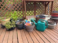 Wide shot of plastic and ceramic pots, gardening gloves, hand tools, watering can, all on wooden deck with lattice background.