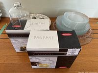 Glass butter dish with dome lid beside two boxes of Rachael Ray Gourmet Glassware butter dish sets, on a wood surface