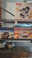 Photo showing T-fal EZ Clean Family deep fryer box, Rival electric food slicer box, Waring Pro meat grinder box, metal saucepans, wok, and oven racks arranged on shelves and countertop.