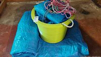 Photo showing blue tarps folded on floor with yellow bucket containing ropes on top