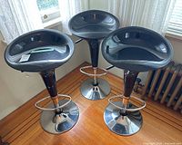 Three black plastic/resin and chrome metal adjustable bar stools grouped together on wood floor near windows.