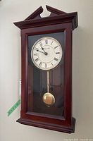 Front view of the wooden pendulum wall clock showing the Roman numeral dial and brass pendulum inside the glass case.