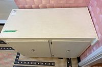 Top front view of a light beige painted wooden cabinet with two doors and silver round knobs on a tiled floor next to a pink tiled wall.