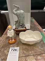 Three items displayed together on marble table: stone heron carving on wooden base, small figurine of child, and Belleek porcelain bowl.