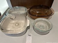 Four pieces of glass cookware including two Corningware and Visionware casserole dishes with lids, and two smaller round glass bowls with lids.