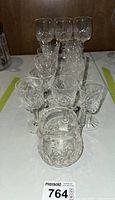 Mixed crystal stemware including small appetizer glasses, wine glasses, and a crystal pitcher displayed on white surface with tablecloth beneath