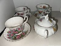 Set of three vintage fine china teacups with matching saucers and an ornate sugar bowl with lid, arranged on a shelf.