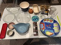Overview of assorted kitchenware including clear glass dishes and bowls, ceramic bowls, decorative plates with fruit and flower motifs, two drinking glasses, a small wooden rack with kitchen tools, and a small terracotta container.