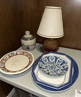 Overview photo of ceramic serveware including decorative plates, bowl, pottery vase, and table lamp on a white surface against wood background.