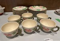 Six ceramic tea cups with a pink flower and green leaf design, cream colored background, gold or brown rim detail, arranged in a row.