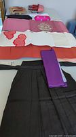 Traditional Japanese attire laid out on a surface showing various fabric pieces including pink, cream with red embroidery, orange textiles, a purple sash, and black hakama pants.