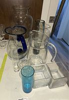 Lot of nine glass items including a large clear vase, pitchers with blue trim, and smaller clear and blue glass vessels arranged on a table.