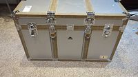 Front view of the silver metal trunk with gold reinforcement strips, central lock clasps, and leather handle.