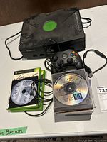 Original Xbox console, controller, and game discs displayed on a white surface. Console and controller show dust and signs of use. Several game discs including Call of Duty 2 and Exite are visible.