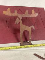 Rustic metal moose sculpture in front of the red metal sign with maple leaf cutout to display size and design.