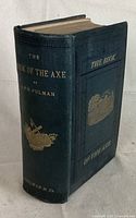 Book standing closed showing spine with gilt title 'THE BOOK OF THE AXE by C.P.R. Pulman' and an emblem of an axe with hands in gold on the spine, dark cloth with embossed pattern.