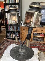 Front view of both antique coach lanterns mounted on forged stand in indoor setting, showing original glass panels and brass tops.