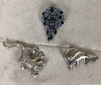 Front view of the two silver animal brooches (gecko and dinosaur) and one blue glass brooch placed on fabric background.
