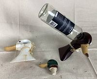 Photo showing white wooden duck wine bottle stand and purple wooden duck wine bottle stand, each holding a wine bottle inverted, alongside the green-headed duck wine stopper.