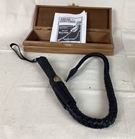 Open wooden box with black braided leather whip laid out beside it and a folded paper inside the box