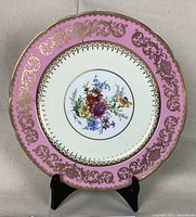Front view of decorative plate showing deep pink border with gold accents and floral center.