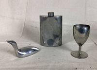 Three pewter items on a flat surface: a Canadian Hoselton loon sculpture, a dented hip flask, and a small wine cup.