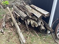 Pile of weathered and aged split rail wooden pieces stacked outdoors on grass and near a shed, showing various lengths and heavy wear.