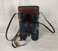 Binoculars with visible flaking finish and attached straps in front of white background, with brown leather case behind.