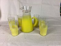 Full set shown: a lemon yellow glass carafe and six matching gradient glasses arranged around it on a neutral fabric background.