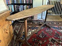 Full angle photo showing the vintage wooden ironing board set on the wooden frame stand on a patterned rug with bookshelves and wood panel walls in the background.