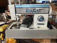 Front and side view of the white and blue Janome Vogue Stitch 5800 sewing machine on its case, with foot pedal visible.