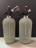 Two vintage soda syphon seltzer bottles with black plastic tops and silver wire mesh, standing side by side against a black backdrop. They measure approximately 14.5 inches tall.