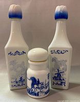 Front view of two white glass Vandermint Dutch windmill liquor bottles with blue poodle and windmill decorations, with blue gradient tulip stoppers, shown alongside a smaller white glass jar with a blue horse and rider design and white stopper.