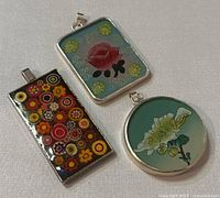 Front side of three costume jewelry pendants with colorful floral designs inside silver frames.