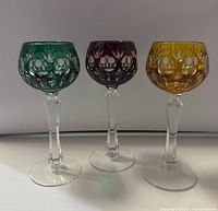 Front view of the three colored crystal wine glasses in green, purple, and amber with detailed cut glass designs, set on white surface.