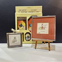 Five small framed oil paintings visible in original packaging, a framed Wanda Lee perfume bottle print, and a framed matted signed cartoon on display.