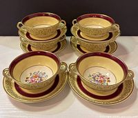 Six pairs of double handle cups and matching saucers arranged in two rows. Cups are cream with maroon and gold floral detail and floral interior.