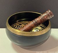 Side angle of the black and brass singing bowl with wooden mallet inside, showing the geometric flower pattern within the bowl.