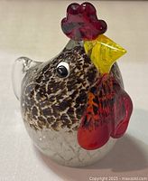 Front view of hand blown glass rooster paperweight showing detailed speckled body, yellow beak, and red comb and wattles.