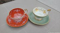 Two Aynsley bone china teacup and saucer sets side by side on a tiled surface, one orange with white floral patterns and gold trim, the other green with gold trim.