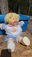 Front view of the Cabbage Patch Kids doll showing facial features, blonde yarn hair with red bow, blue gingham dress, and white bib with giraffe embroidery.