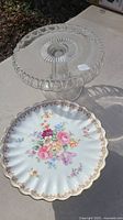 Photo showing the plate with floral design and scalloped gold rim sitting on outdoor surface alongside a glass pedestal stand.